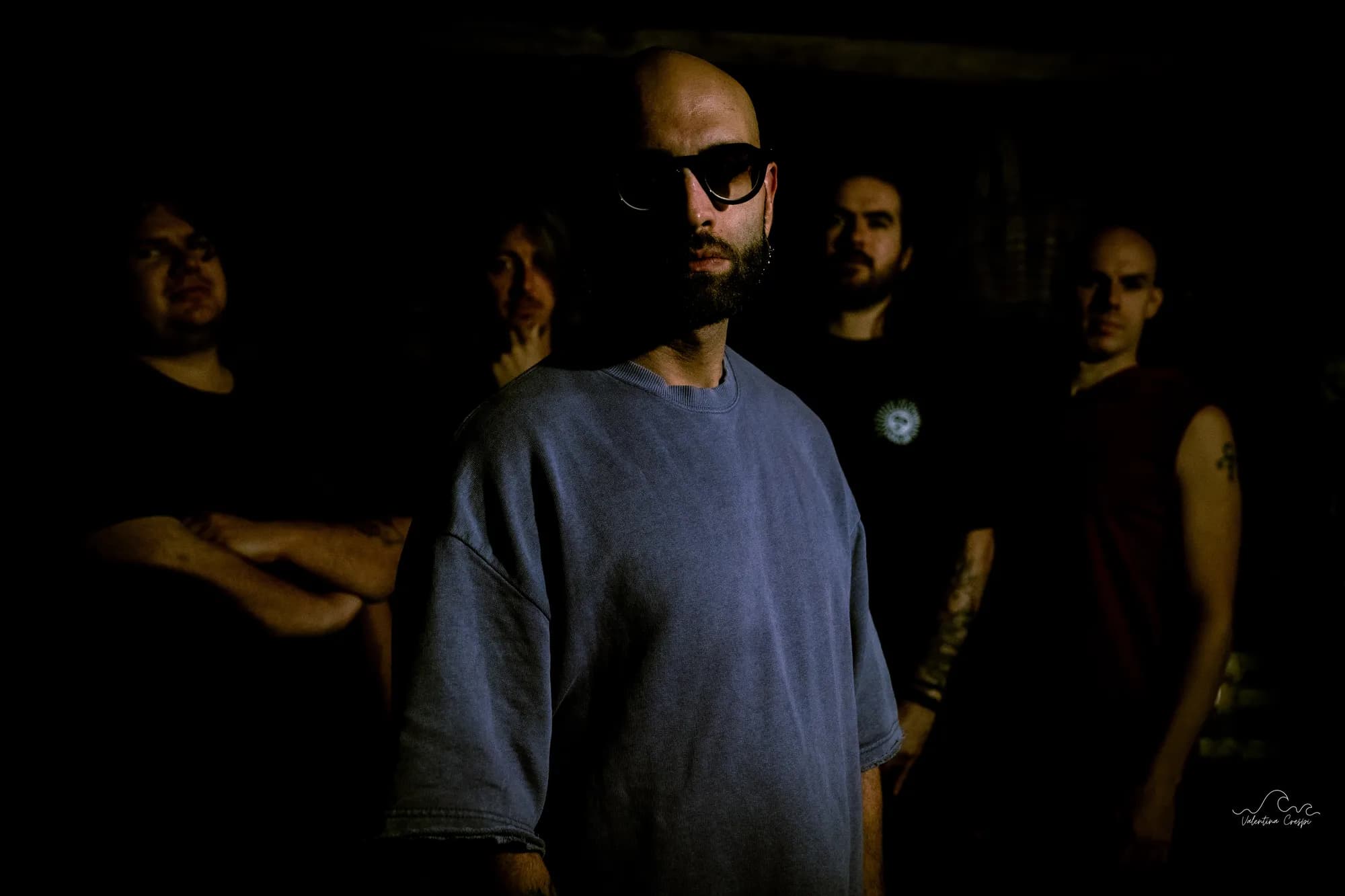 D-DOC in primo piano con occhiali da sole e i Cavalieri del Sacromonte alle spalle, nella penombra