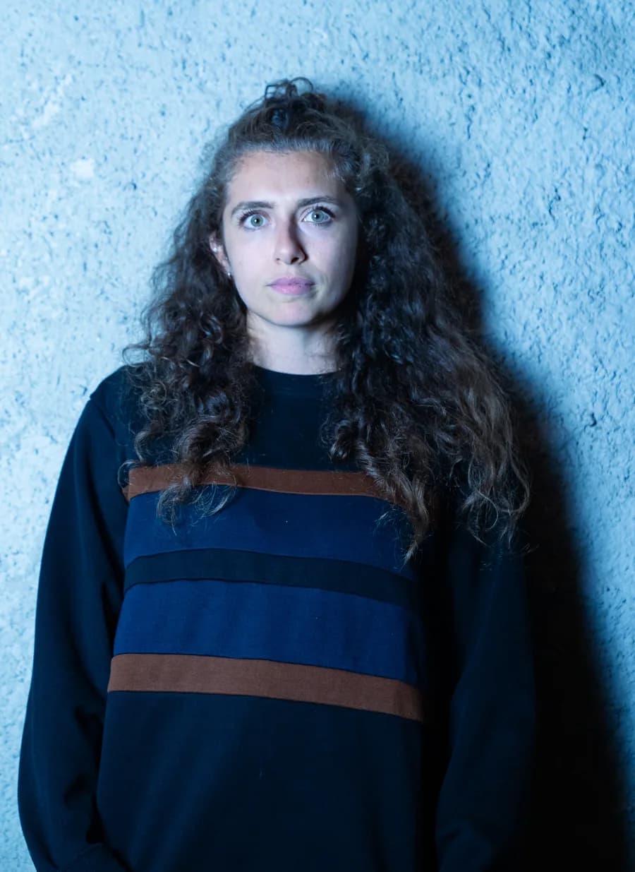 Priscilla con capelli ricci e maglione a righe davanti a un muro illuminato di azzurro