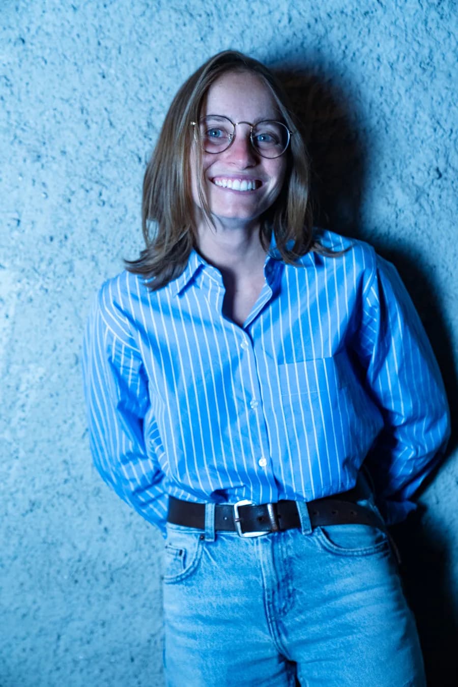 Valentina Crespi sorridente, con occhiali e camicia a righe, davanti a un muro illuminato di azzurro